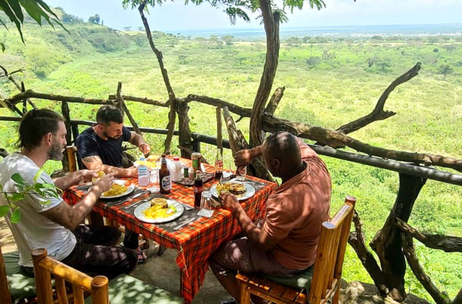 camp-food-for-tourists-uganda - Rent A Driver Uganda Three men dining at a small wooden table on a balcony overlooking a lush green valley and distant horizon.