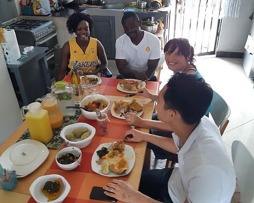 tourists-eat-food-in-kampala - Rent A Driver Uganda Four people sit around a colorful orange table enjoying a meal in a bright kitchen, with plates of food and pitchers of juice nearby.